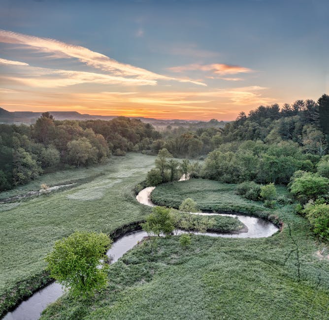 An expansive garden landscape at sunset, featuring a winding river flowing through lush green fields and clusters of mature trees. The river's gentle curves create serene borders around well-maintaine